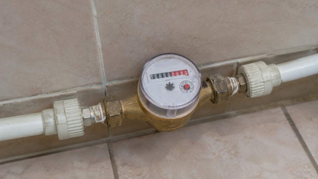 Vodomerno brojilo u kupatilu spojeno na bele cevi pored keramičkih pločica.